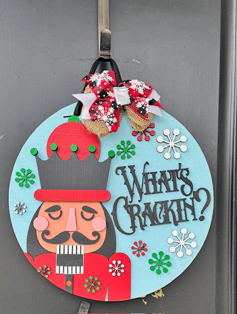 Decorative door hanger with a nutcracker design and text 'What's Crackin?' on a light blue background.
